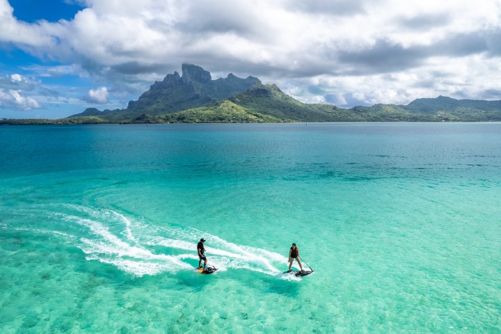 a man riding a wave on top of a body of water with Bora Bora in the background